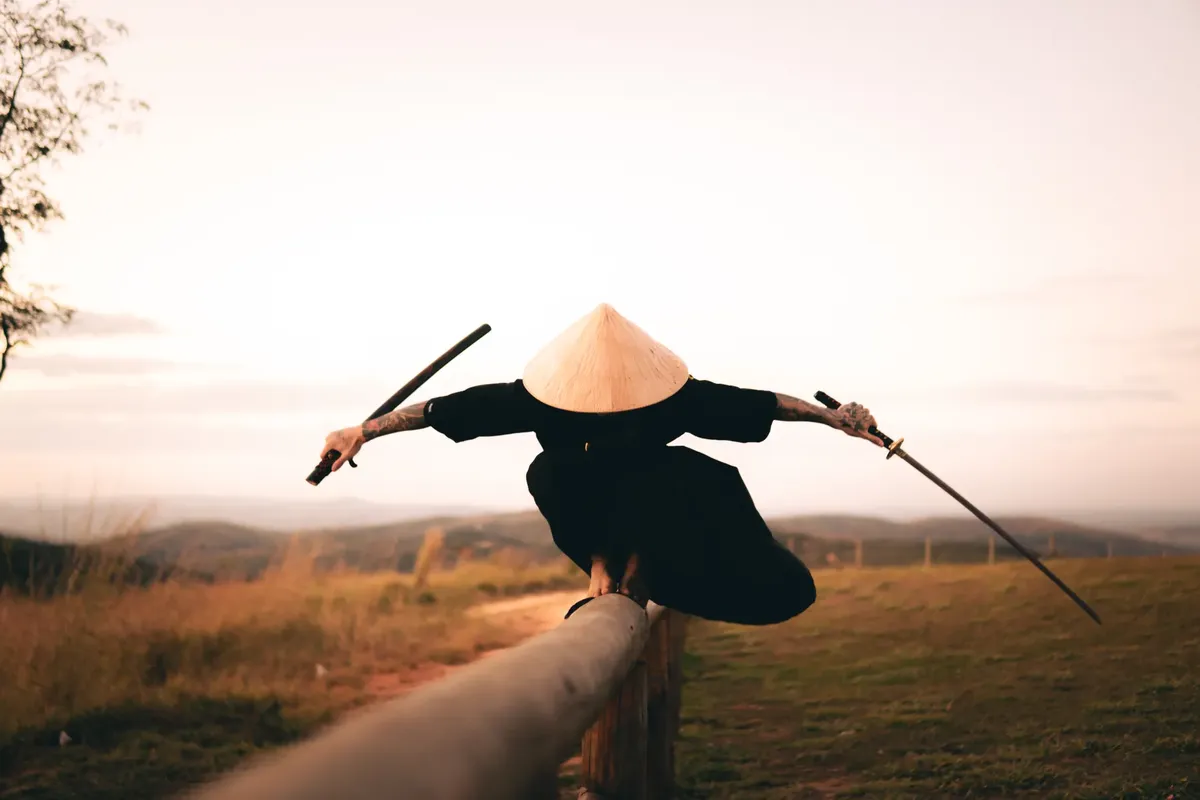 Martial artist in traditional hat balances on a fence wielding samurai swords outdoors at sunset.